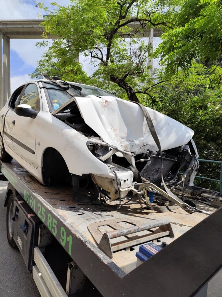 Épave voiture blanche sur dépanneuse après accident Épave de voiture blanche détruite à l'avant, chargée sur une dépanneuse. Viaduc et feuillage vert en arrière-plan. Tél. dépannage : 06 61 25 85 91.